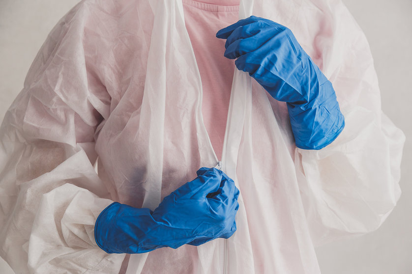 Doctor epidemiologist puts on white protective suit, fastens a zipper, hands close-up. Hands in protective rubber gloves. Personal protective equipment concept.