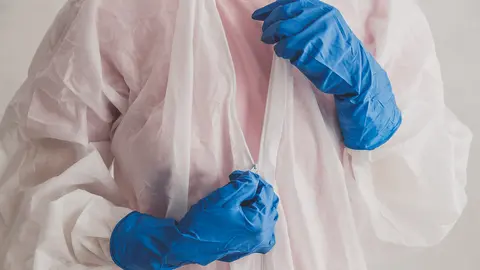 Doctor epidemiologist puts on white protective suit, fastens a zipper, hands close-up. Hands in protective rubber gloves. Personal protective equipment concept.
