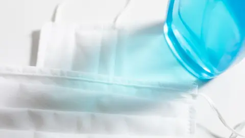 High angle view close up of a sanitizer and two white face masks protection against pandemic coronavirus covid 19 and for safety, lying on a white table 